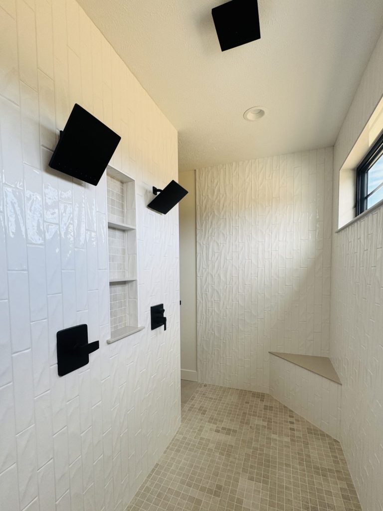 Modern walk-in shower with white textured tile walls, built-in shelving, a corner bench, and matte black fixtures, lit by natural light from a window.