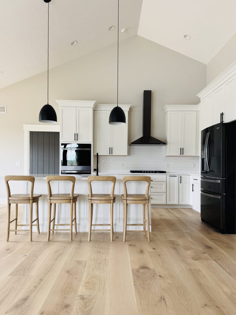 Modern kitchen with light wood floors, white cabinets, black appliances, a white island with four wooden chairs, and two black pendant lights hanging above.
