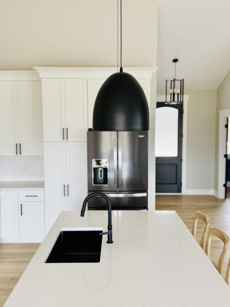 Modern kitchen with white cabinets, a black refrigerator, a large black pendant light, a white island, and a black sink and faucet. Two wooden chairs are at the island.