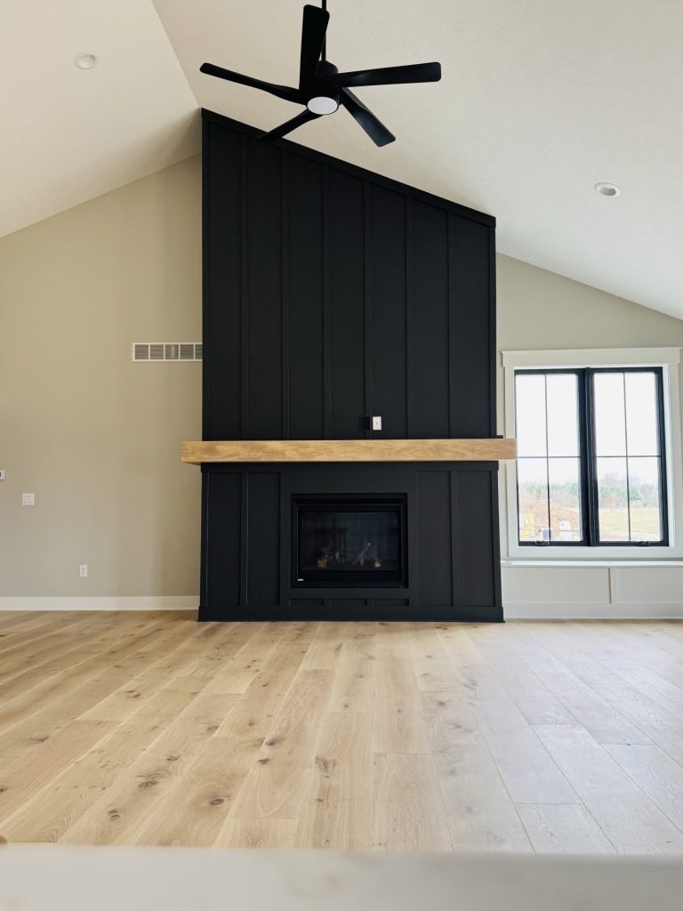 A modern living room with light wood flooring, a black paneled fireplace wall, a wooden mantel, ceiling fan, and a large window.