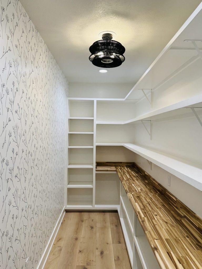 A walk-in pantry with white shelving, wood countertops, open shelves, a patterned wallpaper accent wall, and a modern ceiling light fixture.
