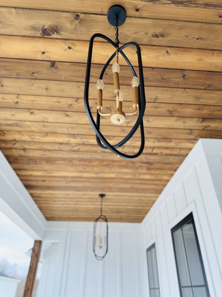 A modern pendant light with a black geometric frame and exposed bulbs hangs from a wooden ceiling on a covered porch. A second, similar light is visible in the background.