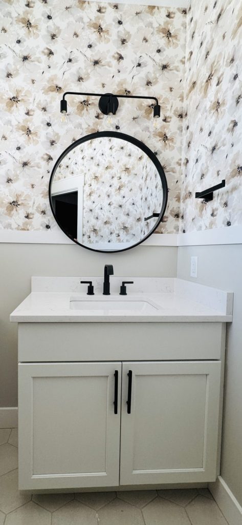 A small bathroom vanity with a white countertop, black faucet, round mirror, and floral-patterned wallpaper. Black light fixture and towel holder are mounted on the wall.