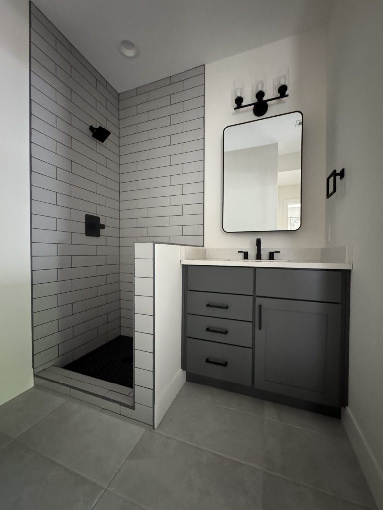 A modern bathroom with a corner walk-in shower, gray subway tiles, a black shower fixture, gray vanity with black hardware, a rectangular mirror, and wall-mounted light above.