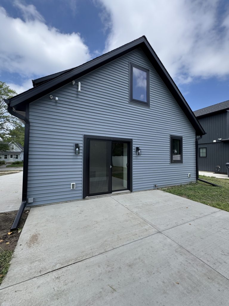 A modern two-story house with blue-gray siding, black trim, a large upper window, sliding glass doors, and a concrete patio.