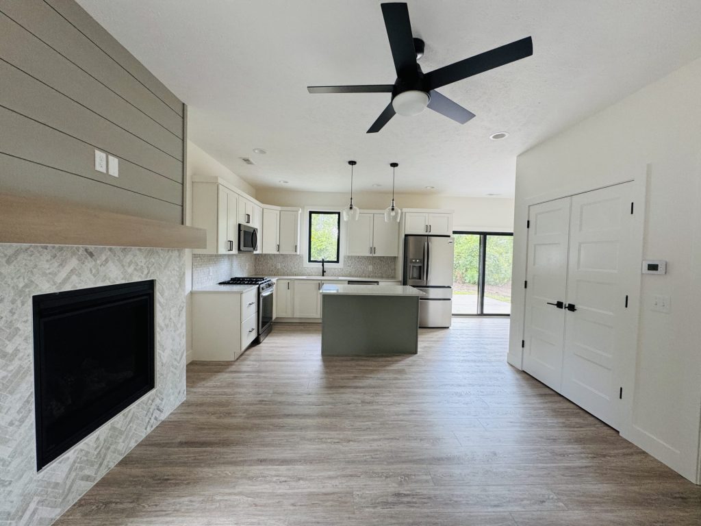 Modern kitchen with white cabinets, a center island, stainless steel appliances, wood flooring, a ceiling fan, a fireplace, double doors, and large windows providing natural light.