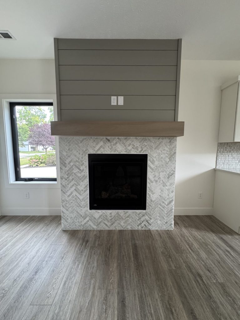 Modern living room with a fireplace featuring a herringbone tile surround, wood mantel, vertical shiplap paneling above, and wood-look flooring. Large window on the left.