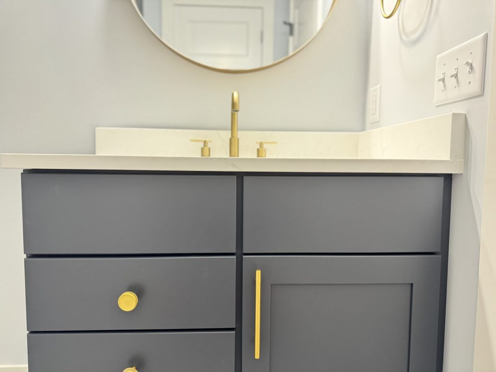 Gray bathroom vanity with gold handles and faucet, white countertop, round mirror, and light switches on the wall.