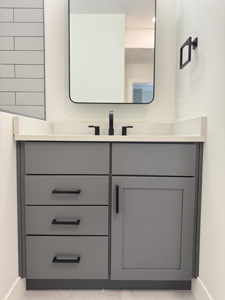 A modern bathroom vanity with a gray cabinet, black hardware, white countertop, black faucet, rectangular mirror, and a towel holder on the wall.
