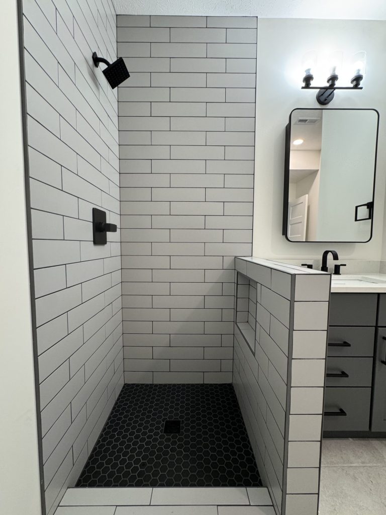 Modern bathroom shower with large light gray subway tiles, black fixtures, hexagonal black shower floor tiles, a built-in niche, and a vanity with a rectangular mirror and lights.