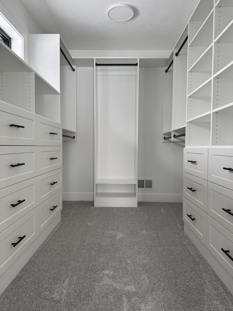 A modern, empty walk-in closet with white built-in shelves, drawers, hanging rods, and grey carpeted floor.