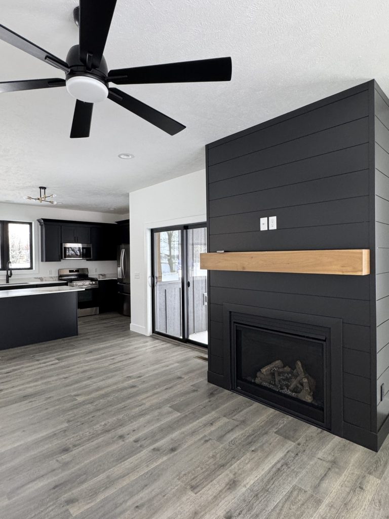 Modern living space with a black shiplap fireplace and wood mantel, adjacent to a kitchen with black cabinets and large ceiling fans. Sliding glass doors open to an outdoor area.