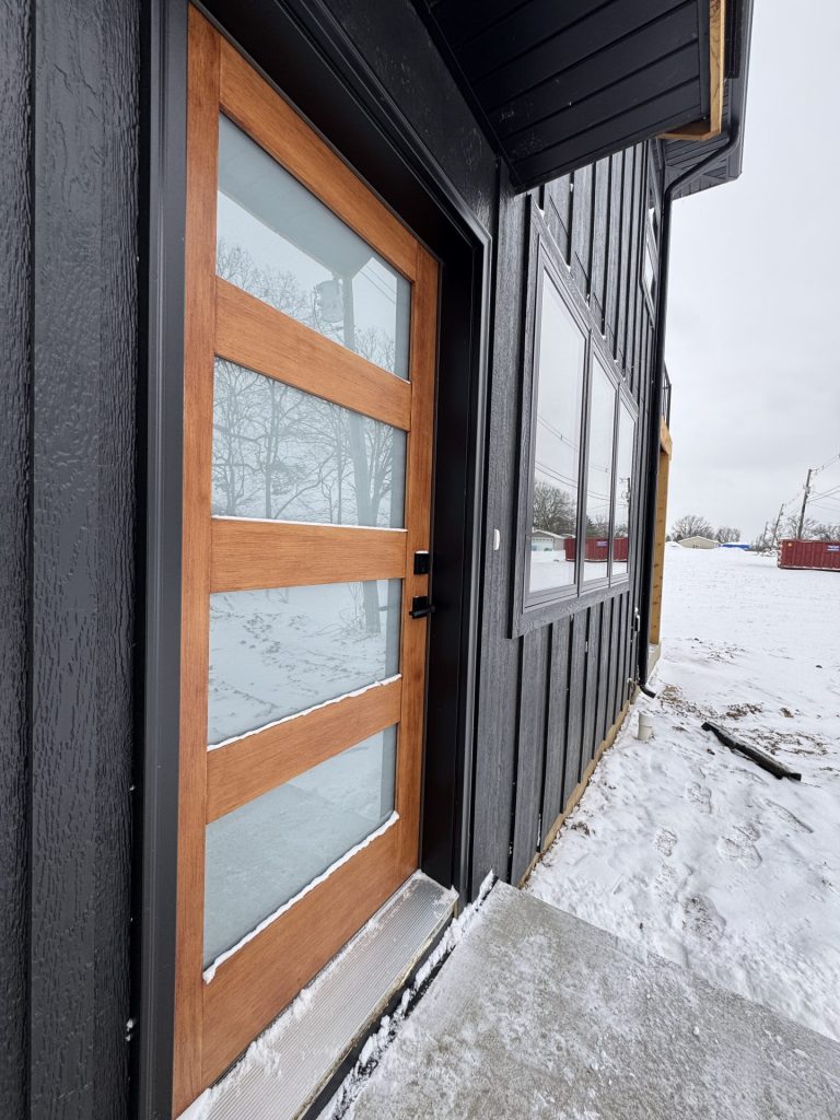 A modern wooden and glass front door on a black house, with snow on the ground and bare trees in the background.
