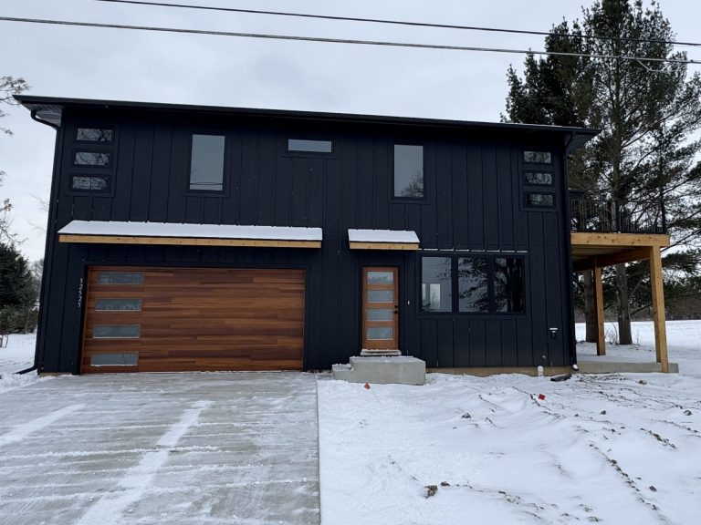 A modern two-story black house with wood accents, large windows, and a snow-covered driveway and yard.