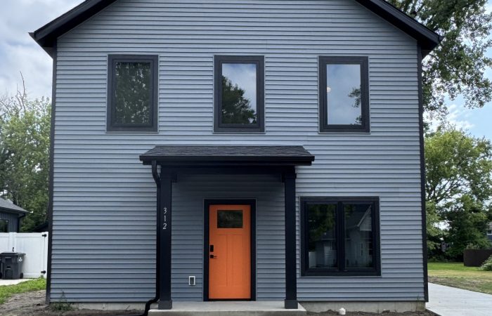 Two-story modern house with gray siding, black trim, and an orange front door, featuring a small porch and a concrete walkway, surrounded by bare soil and trees.
