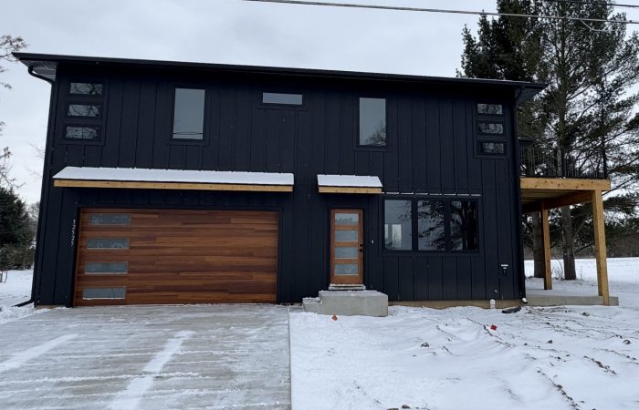 A modern two-story black house with wood accents, large windows, and a snow-covered driveway and yard.